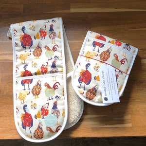 May include: A pair of white double oven gloves with a colourful chicken print. The gloves are made from 100% cotton panama and have a heat-resistant towelling lining. The text on the tag reads "Double Oven Gloves", "Made from 100% Cotton Panama", "Lining is made from heat resistant towelling", "Made in the UK".
