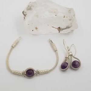 May include: A silver bracelet and earrings set featuring amethyst stones. The bracelet is made from a braided silver chain and has a single amethyst stone in the centre. The earrings are also made from braided silver chains and each has a single amethyst stone.