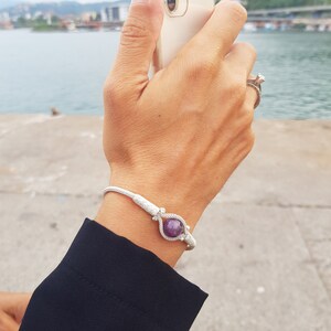 May include: A white braided bracelet with a silver clasp and a purple gemstone. The bracelet is on a person's wrist.