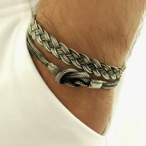 May include: Two silver bracelets. The top bracelet has a thick, braided design. The bottom bracelet is thinner, with a knotted closure. Both bracelets are worn on a wrist, against a white shirt.