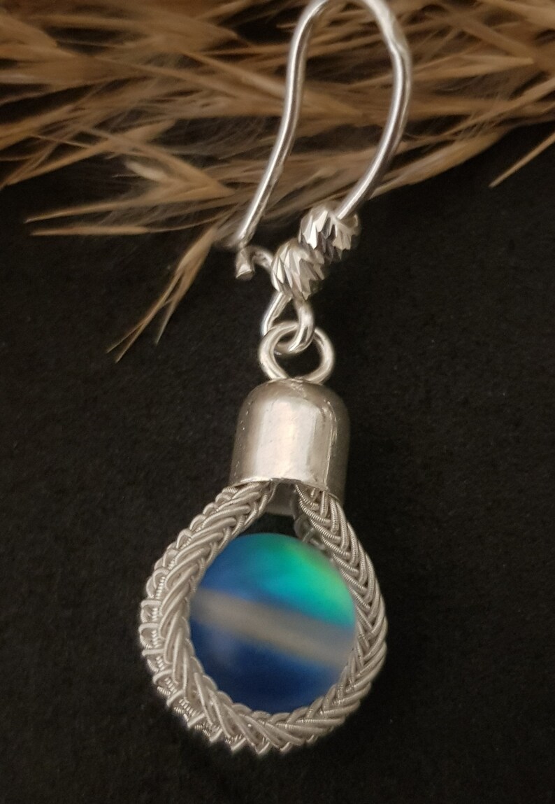 May include: Silver earring featuring a blue, green and white iridescent glass bead enclosed in a silver wire cage.