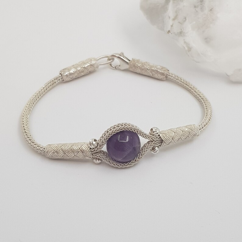 May include: A silver bracelet with a braided chain and a single round amethyst gemstone.