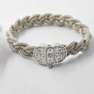 May include: A silver braided bracelet with a decorative clasp. The clasp is made of two oval shapes with intricate floral designs.