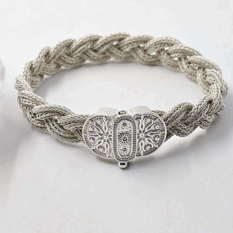May include: A silver braided bracelet with a decorative clasp. The clasp is made of two oval shapes with intricate floral designs.
