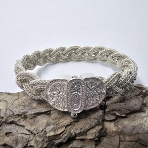 May include: A silver bracelet with a braided design and a decorative clasp. The clasp is detailed with a floral pattern.