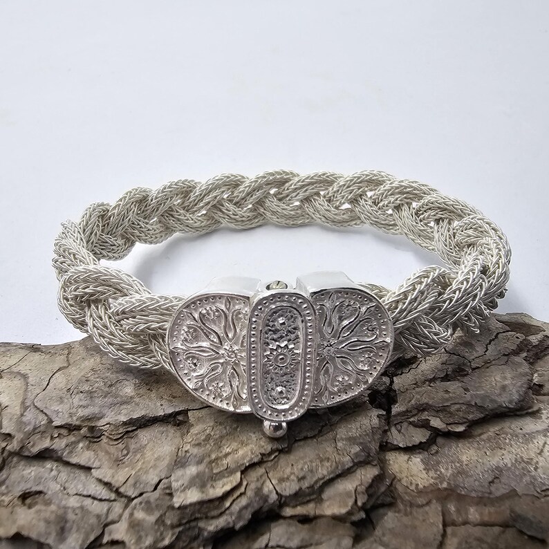 May include: A silver bracelet with a braided design and a decorative clasp. The clasp is detailed with a floral pattern.