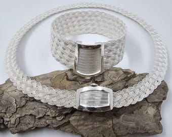 Puede incluir: Un conjunto de collar y pulsera de plata con un diseño trenzado y un cierre rectangular. Las joyas se muestran sobre una pieza de madera desgastada.