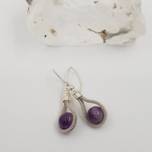 May include: A pair of silver earrings with a purple amethyst stone in each. The earrings are made with a woven wire design.
