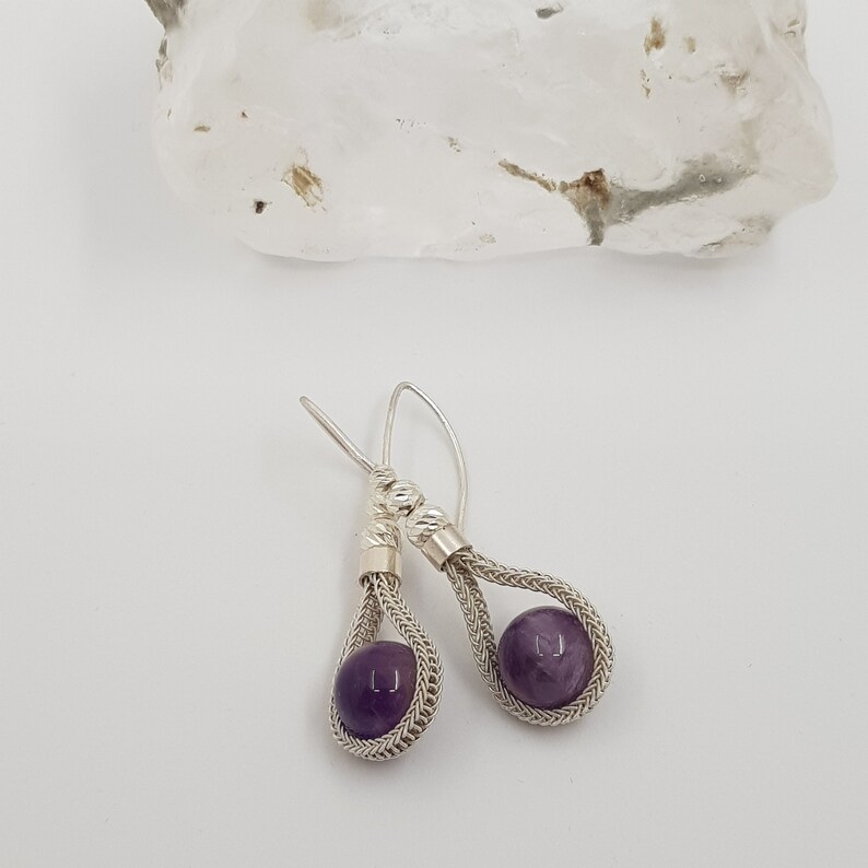 May include: A pair of silver earrings with a purple amethyst stone in each. The earrings are made with a woven wire design.