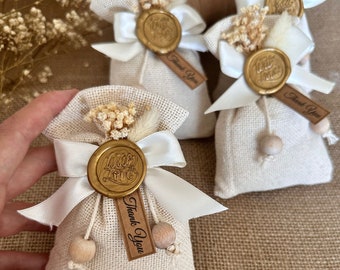 May include: Three small white burlap bags with white satin ribbon bows and gold wax seals. Each bag has a small brown tag that says "Thank You". The bags are filled with dried white flowers.