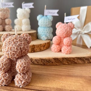 May include: A selection of teddy bear-shaped candles in shades of brown, white, blue, and pink. Each candle features a rose design. Some candles have small flags that read "Thank you". The candles are presented on wooden slices, with a gift box in the background.