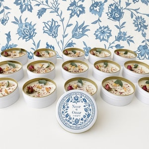 May include: White candles in round tins with gold rims, decorated with dried flowers and herbs. The candles are arranged on a white surface, with a blue and white floral wallpaper background. One candle lid is visible, with the text "Noor & Omar".