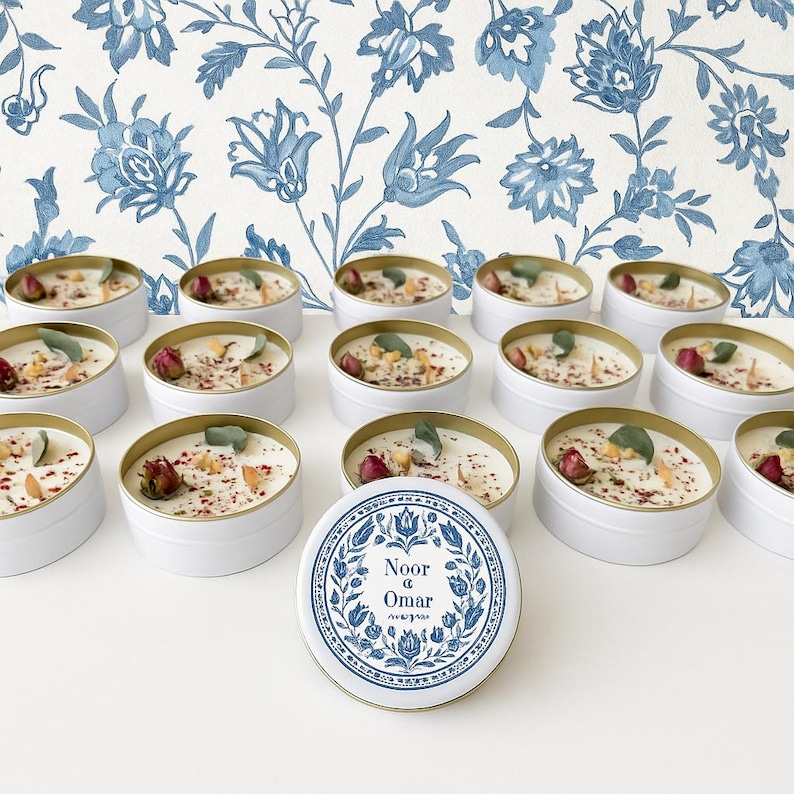 May include: White candles in round tins with gold rims, decorated with dried flowers and herbs. The candles are arranged on a white surface, with a blue and white floral wallpaper background. One candle lid is visible, with the text "Noor & Omar".