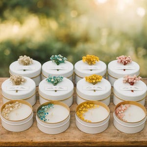 May include: A collection of white candles in tins with gold rims, adorned with dried flowers in shades of beige, blue, yellow, and pink. Each candle has a label that reads "Let it Shine". The candles are on a wooden surface.