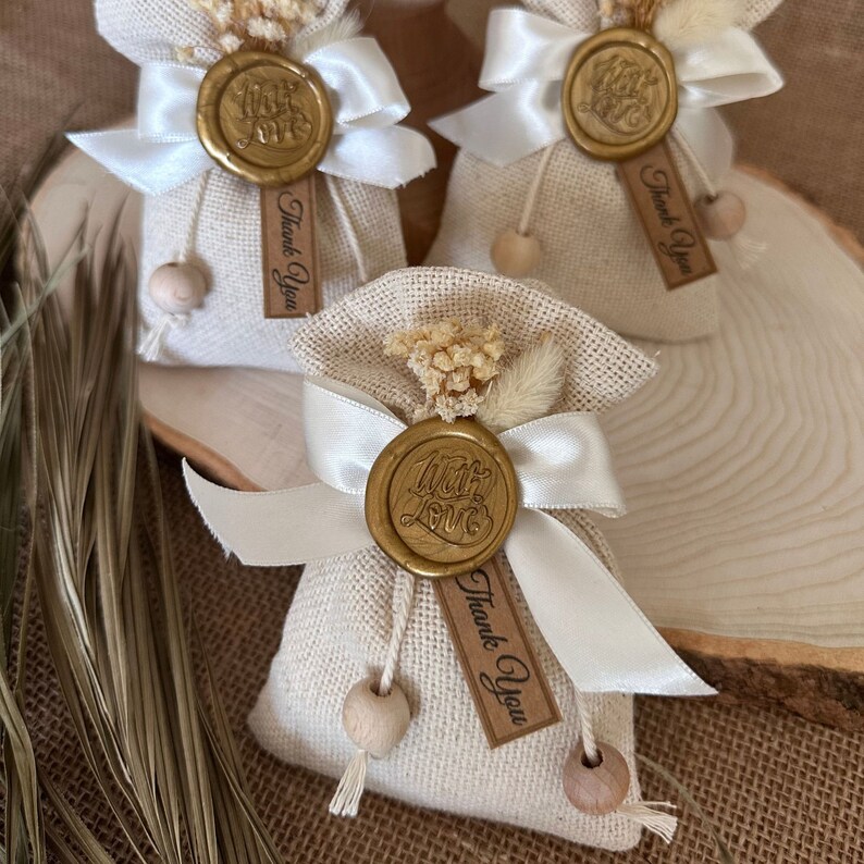 May include: Three small, beige burlap bags with white ribbon bows and gold wax seals that say "With Love". Each bag has a small wooden bead and a brown tag that says "Thank You".