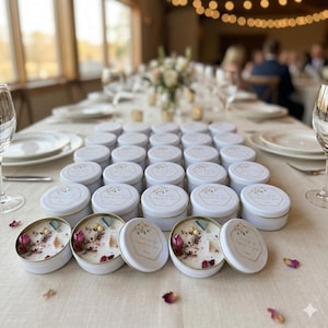 Puede incluir: Pequeñas velas redondas blancas en latas con bordes y tapas doradas. Las tapas tienen un diseño floral y texto. Algunas latas están abiertas, revelando la cera, flores secas y cristales. Las velas están dispuestas sobre una mesa.