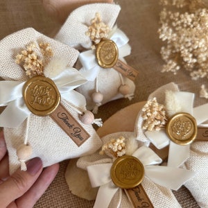 May include: Set of four small, beige burlap bags with white satin ribbon ties. Each bag has a gold wax seal with the words "With Love" and a small wooden bead. The bags are decorated with dried white flowers and a small tag that says "Thank You".