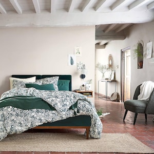 May include: A green velvet bed with a white and green floral duvet cover and a green throw pillow. The bed is in a bedroom with a white ceiling and white walls. There is a wooden bedside table next to the bed with a lamp and a vase of flowers.