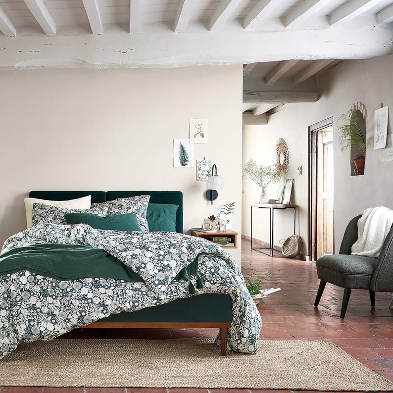 May include: A green velvet bed with a white and green floral duvet cover and a green throw pillow. The bed is in a bedroom with a white ceiling and white walls. There is a wooden bedside table next to the bed with a lamp and a vase of flowers.