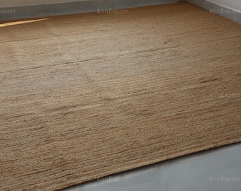 May include: A rectangular area rug made of natural tan jute fibers, featuring a tightly woven, flat-weave construction with a visible horizontal grain pattern. The rug has a consistent, earthy brown tone throughout its surface and a clean, straight edge.