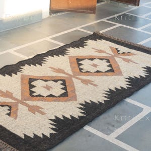 May include: Rectangular rug with a geometric design. The rug features a cream-colored central field with brown and black diamond and arrow patterns. The border is black with a zigzag pattern, and the rug has a fringed edge.