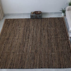 May include: A large, rectangular brown jute rug with a textured, striped pattern. The rug is placed on a light-coloured floor, and is surrounded by a bed, a small chest, and curtains.