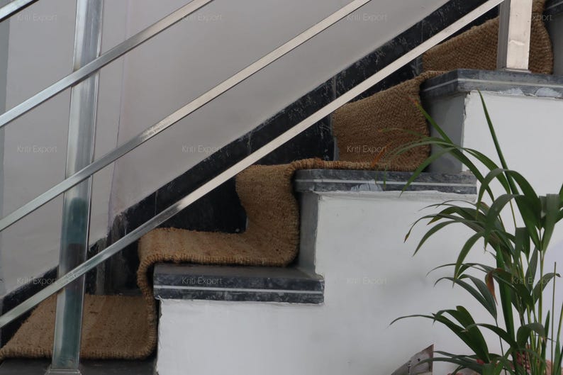 K&ouml;nnte beinhalten: Eine Treppe mit braunen gewebten Stufen. Die Treppe hat eine schwarz-graue Umrandung und ein silbernes Metallgel&auml;nder. Eine gr&uuml;ne Pflanze befindet sich im Vordergrund. Die Worte "Kriti Expert" sind auf den Stufen zu sehen.