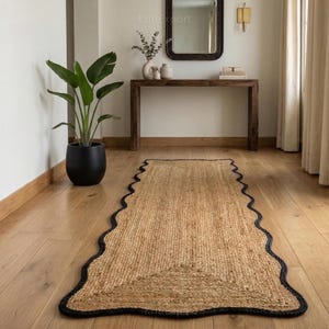 Pode incluir: Um tapete retangular de juta com borda ondulada e borda preta. O tapete é colocado em um piso de madeira. Uma grande planta em vaso e uma mesa de console de madeira estão ao fundo.