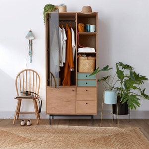 May include: A wooden cabinet with glass doors and drawers. The cabinet is open and has clothes hanging inside. There is a hat on top of the cabinet and a plant in a blue pot on the right side of the image.