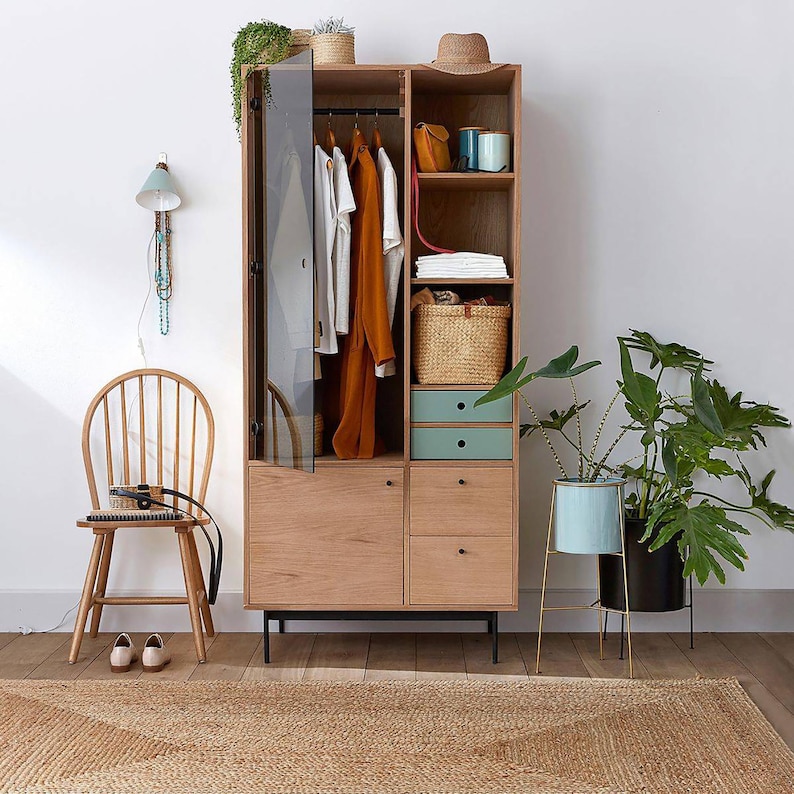 May include: A wooden cabinet with glass doors and drawers. The cabinet is open and has clothes hanging inside. There is a hat on top of the cabinet and a plant in a blue pot on the right side of the image.
