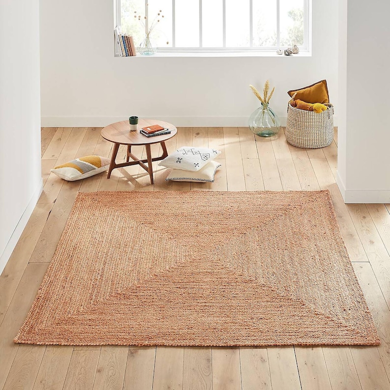 May include: A natural fiber rug with a square design in a light brown color. The rug has a woven texture and a subtle pattern.