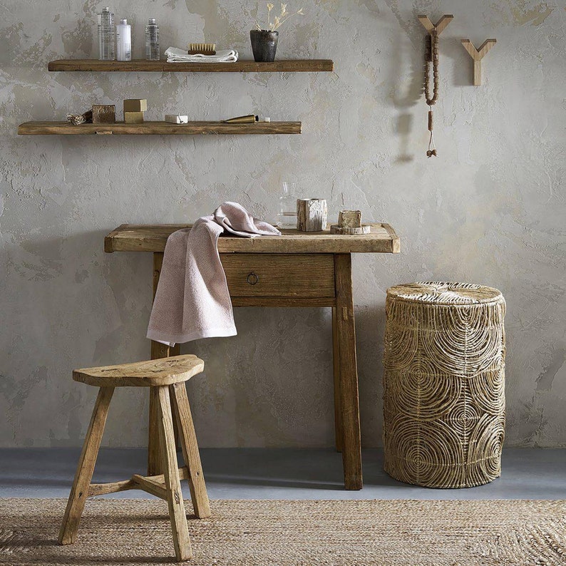 K&ouml;nnte beinhalten: Ein rustikaler Holztisch mit einer Schublade und einem geflochtenen Hocker. Der Tisch hat ein rosa Handtuch dar&uuml;ber gelegt und einen Holzhocker mit einer geschwungenen Sitzfl&auml;che. Tisch und Hocker stehen vor einer Wand mit zwei Holzregalen und einem dekorativen Holz-Wandhaken.