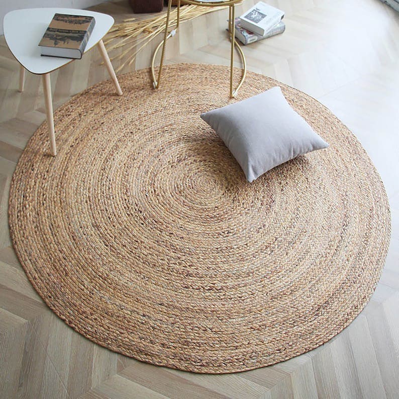 May include: A round, natural fiber rug with a woven texture. The rug is light brown in color and has a circular shape. A light gray throw pillow is placed on the rug.