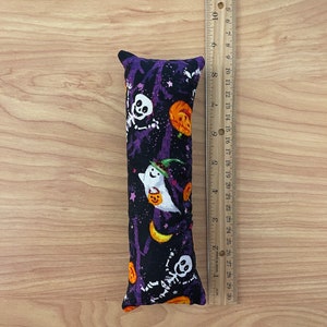 May include: A rectangular cat toy with a Halloween-themed pattern. The fabric features white skeletons, ghosts, and orange pumpkins on a black background with purple tree silhouettes. The toy is approximately 8 inches long.