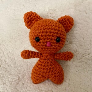 May include: A handmade orange crochet bear with black eyes and a pink nose. The bear has small arms and ears, and a rounded body. The toy is made of a textured yarn and sits on a white, fluffy surface.