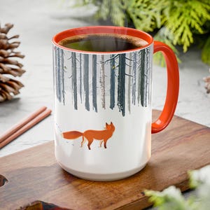 Peut inclure: Une tasse en céramique blanche avec un bord et une poignée orange. La tasse présente une scène de forêt d'hiver avec un renard marchant dans la neige. La tasse est posée sur une planche à découper en bois.