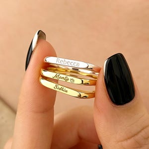 May include: A stack of three personalized rings. The top ring is silver with the name "Rebecca" engraved. The middle ring is gold with the name "Monty" and a small paw print. The bottom ring is gold with the name "Sophia".