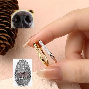 Op de afbeelding: Drie ringen worden tussen de vingers gehouden. Eén is goudkleurig met een vingerafdrukontwerp, een andere is goudkleurig en de derde is roségoudkleurig met een rij kleine, heldere stenen. De afbeelding bevat ook een hondenneus en een vingerafdruk.