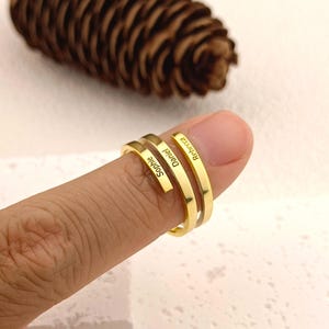 Peut inclure: Une bague dorée à trois bandes, chacune gravée d'un nom : Sophie, Daniel et Rebecca. La bague est portée au doigt, avec une pomme de pin en arrière-plan. La bague est un bijou personnalisé.