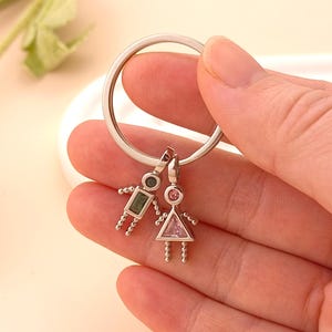 Puede incluir: Llavero plateado con dos colgantes. Un colgante es una figura de niño con una gema rectangular verde y una gema redonda. El otro colgante es una figura de niña con una gema triangular rosa y una gema redonda. El llavero se sostiene en una mano.