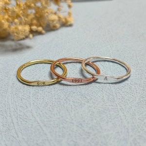 May include: Three thin gold, rose gold, and silver rings with personalized engravings. The gold ring says "SOK" with a heart symbol, the rose gold ring says "1991", and the silver ring says "A".