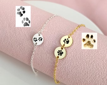May include: A gold chain bracelet with two round charms featuring paw prints. The bracelet is on a pink surface.
