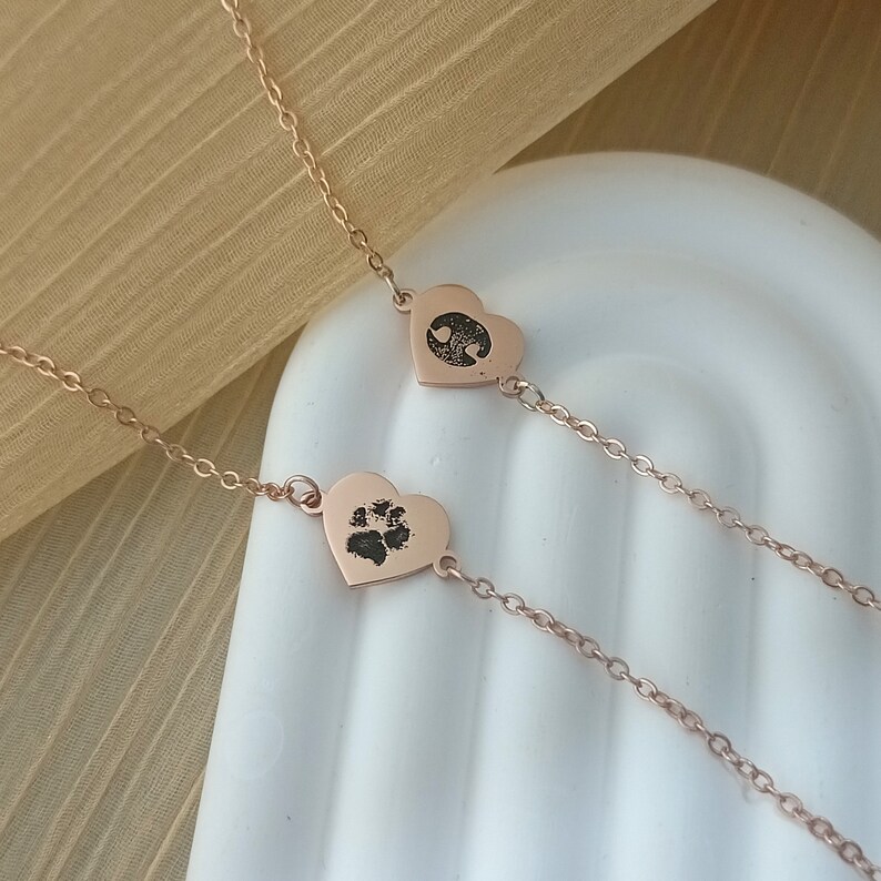 May include: A rose gold necklace with two heart-shaped pendants. One pendant features a paw print and the other has a heart design.