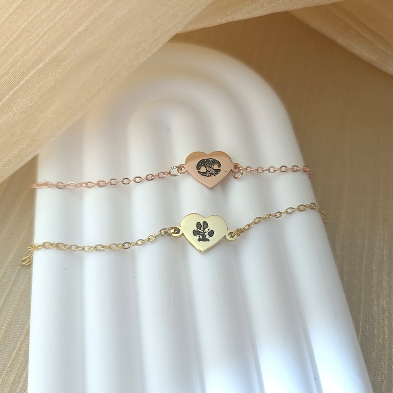 May include: Two delicate heart-shaped bracelets with chain links. One bracelet is rose gold with a tree silhouette inside the heart. The other bracelet is gold with a paw print inside the heart.