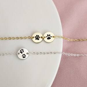 May include: Two delicate necklaces with paw print charms. One necklace has a gold chain and two charms, the other has a silver chain and one charm.