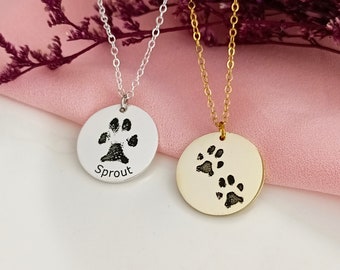 May include: Two silver and gold necklaces with paw print pendants. The silver pendant has a single paw print and the text "Sprout" engraved on it. The gold pendant has two paw prints.