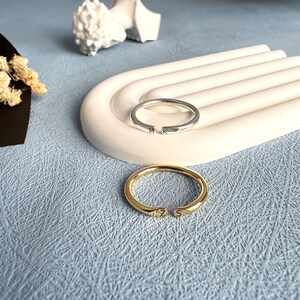 May include: Two simple silver and gold rings with initials engraved on them. The rings are on a white ceramic surface.