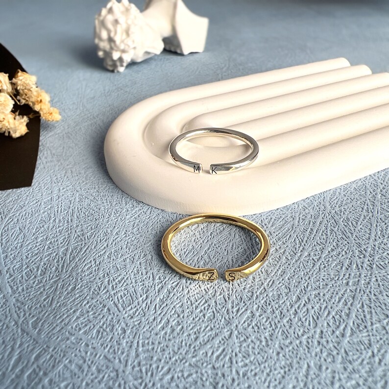 May include: Two simple silver and gold rings with initials engraved on them. The rings are on a white ceramic surface.