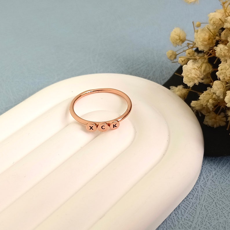 May include: A rose gold ring with three round beads, each with a letter engraved on it. The letters are X, C, and K.