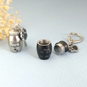 Puede incluir: Dos urnas de cremación plateadas con dijes de huella de pata. Una urna está grabada con "Forever in my heart" y la otra está grabada con "Molly 08.18.20 09.20.20".